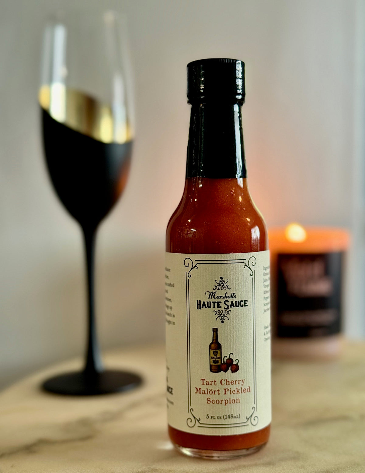 Tart Cherry Malört Hot Sauce bottle next to a wine glass and candle, showcasing bold flavor and spice.