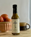 Bottle of Hatch Fermented Leek Scape Matcha Tea Hot Sauce with fresh eggs in the background.
