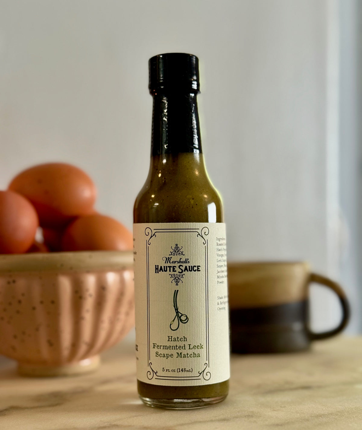 Bottle of Hatch Fermented Leek Scape Matcha Tea Hot Sauce with fresh eggs in the background.